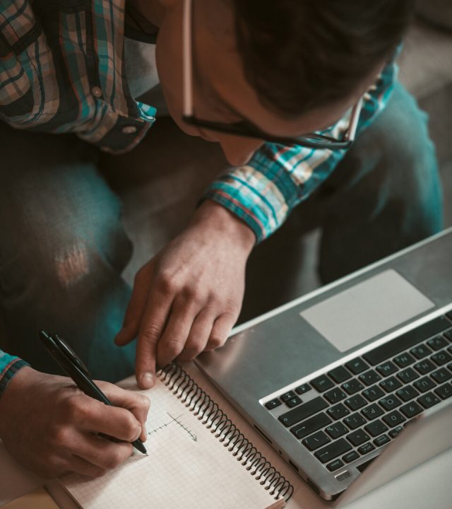 Freelance business concept. Young man writing in notebook at working place. Male freelancer works from home. Close up view from above Freelance business concept. Young man writing in notebook at working place. Male freelancer works from home. Close up view from above.